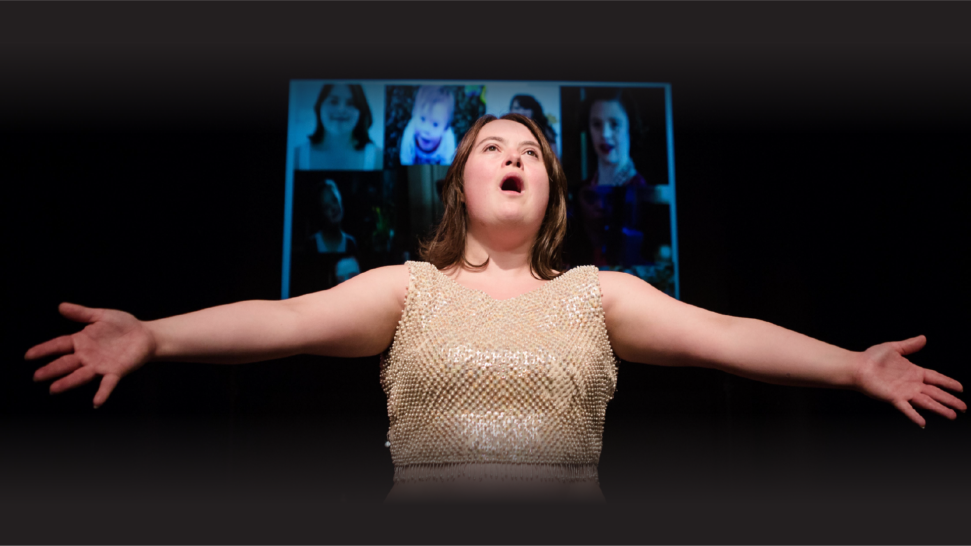A Diploma Student stand in a dark place. She wears a cream color sequin crop top. She has mid-lenght dark blonde haire. She has her arm spread and her mouth is open as if she was singing. Behind her, projected, a mosaic of muliple portraits of people.