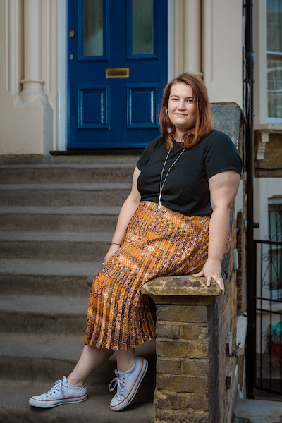 A white woman in a navy top and orange patterned skirt sits on the steps outside a house
