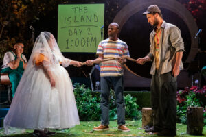 Three members of the company of Imposter 22 stand in the middle of a stage in a pool of light. A large grey hexagon hangs above their heads. They are surrounded by plants and microphones and two other members of the company sitting in the back. A white woman is wearing a wedding gown and a veil over her face. A white man is wearing a grey cap, a grey shirt, a yellow t-shirt and loose dark grey trousers. Between them, a black man wearing a striped t-shirt and blue jeans is having them join hands.