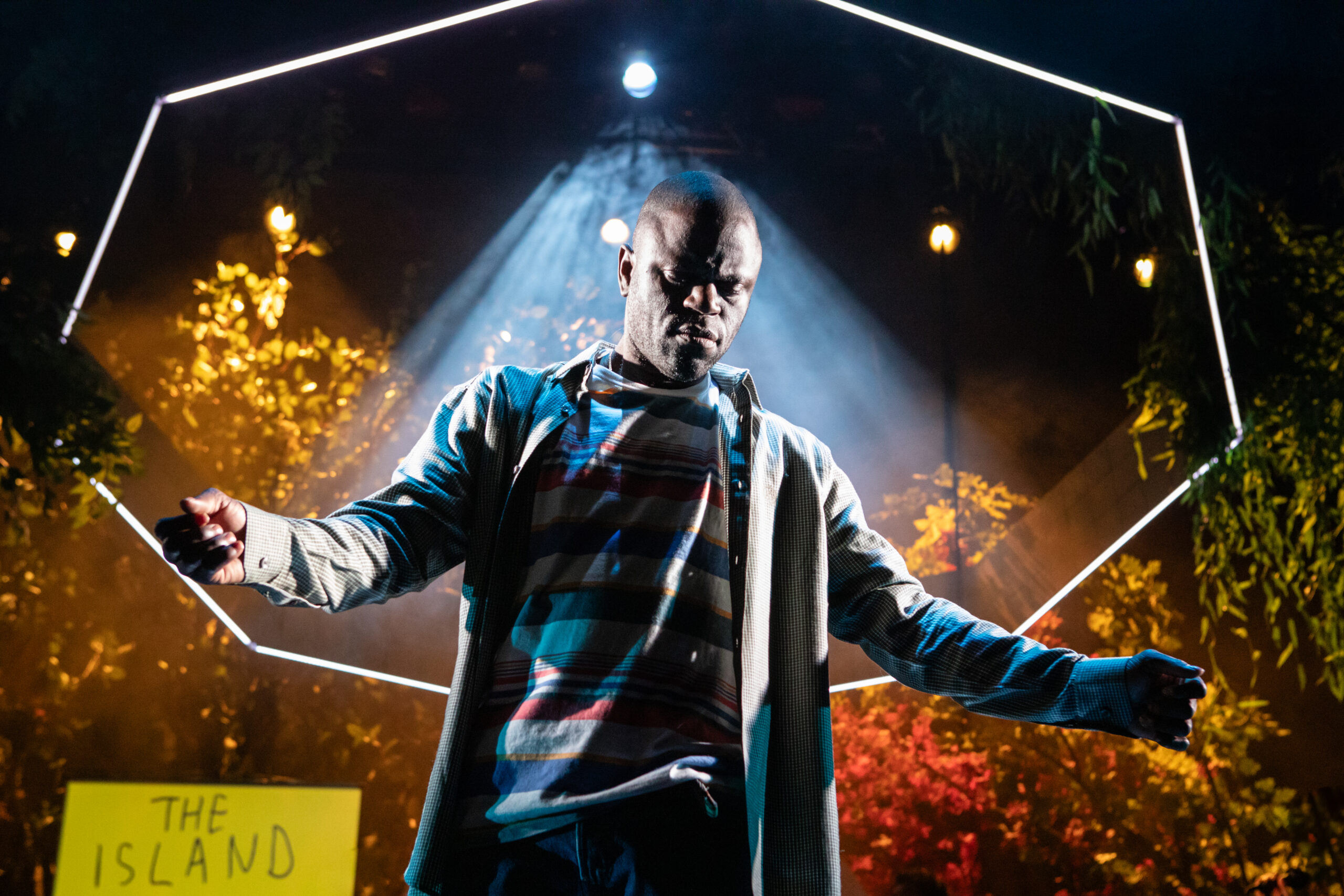 A black man stands in the middle of a stage in a pool of light with his eyes closed and his arm spread. A large white neon hexagon hangs above his head.