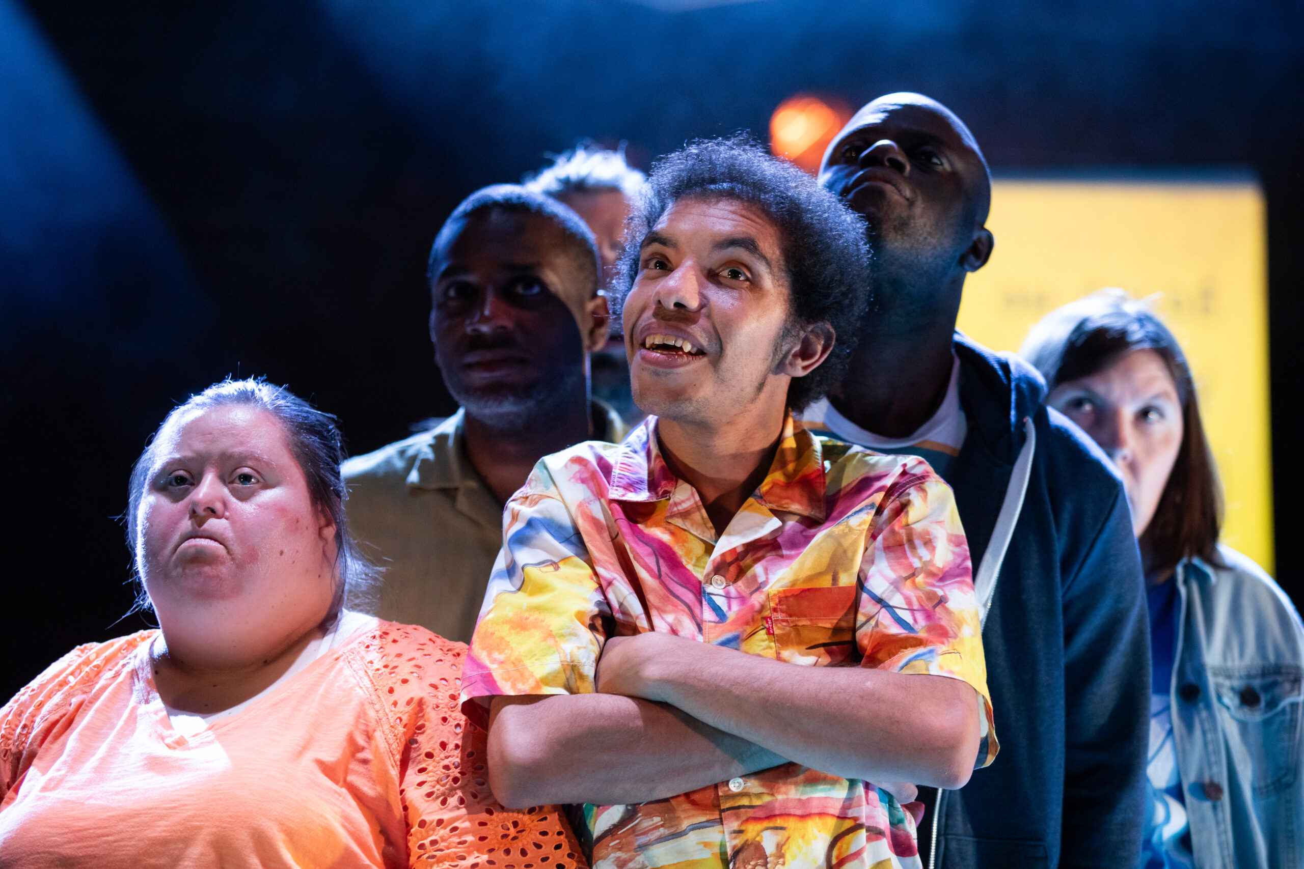 The company of Imposter 22 stands on stage in a pool of blue light. At the front are a black man with dark curly hair and a woman with Down syndrome with dark blond hair tied at the back. The man wears a colourful short-sleeved shirt and has his arm crossed on his chest; he smiles looking ahead with wide eyes. The woman is looking up with a pouting expression. She wears an orange shirt. Behind them stand two black men looking up.