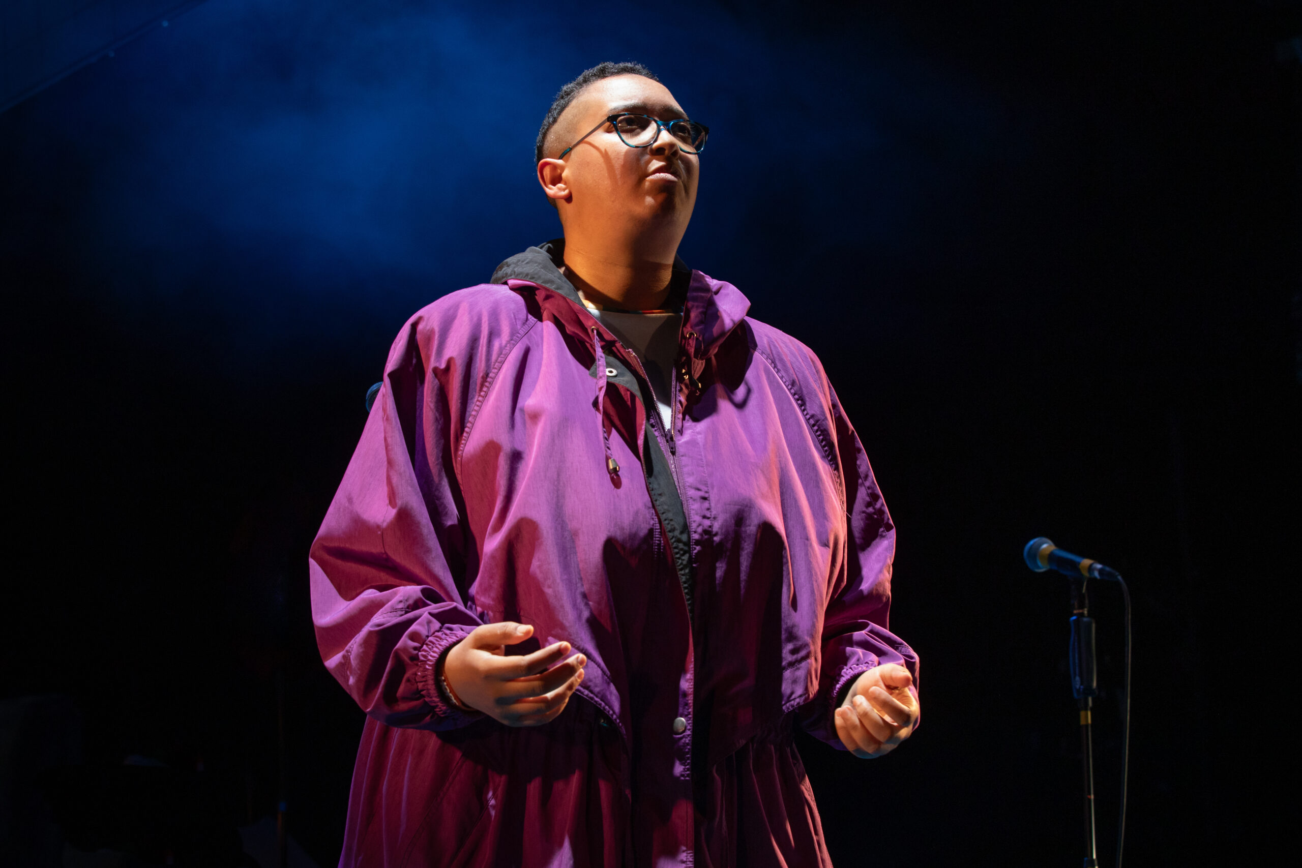 A black woman in a purple raincoat stands in a spotlight. Her hands are out in front of her, palms facing upwards. She has a serious, determined expression on her face.