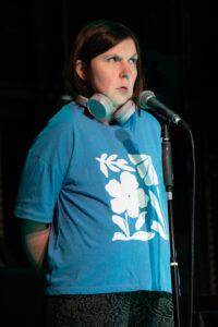 A white woman with dark brown shoulder-length hair, a blue t-shirt with a white floral design stands in a dark space behind a microphone, her hands joined behind her back. She has a pair of grey headpho﻿nes around her neck. She as a dubious look on her face.